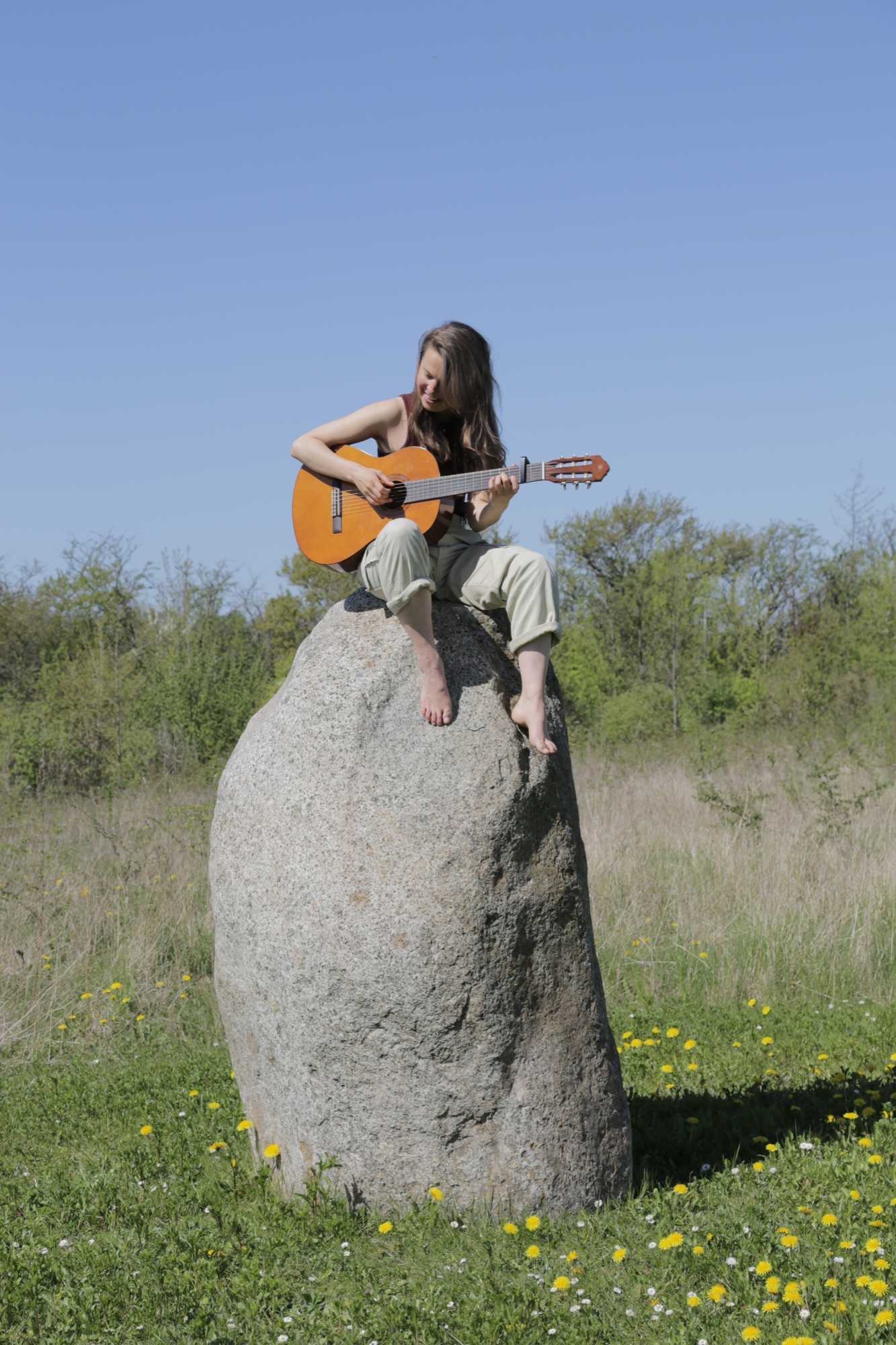 Emma van den Boogert sitting on a standing stone in a field with her guitar